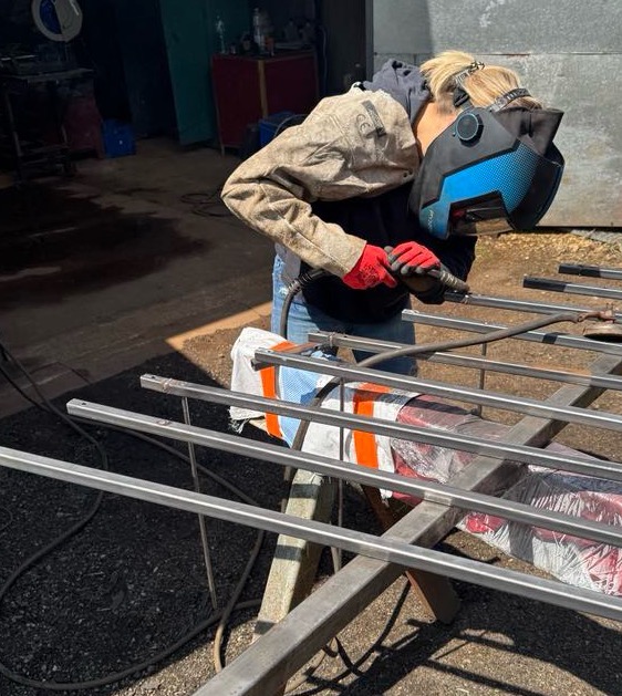 Spawaczka w masce i rękawicach spawa metalową konstrukcję w warsztacie. Widoczne iskry i elementy konstrukcyjne. Ujęcie z boku, w tle warsztatowe wyposażenie.