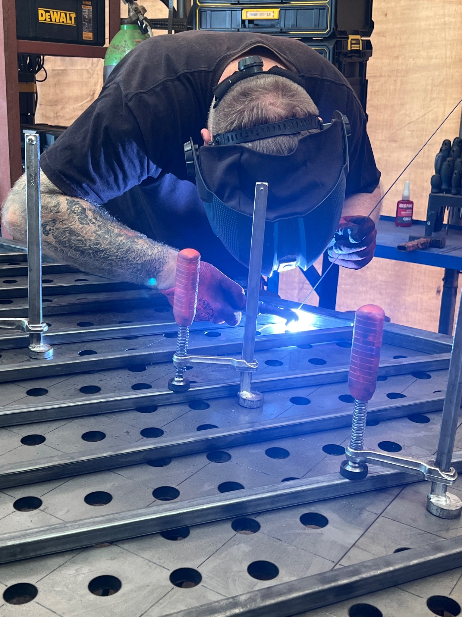 Spawacz w masce spawalniczej łączy metalowe elementy na perforowanym stole spawalniczym, używając zacisków z czerwonymi uchwytami.