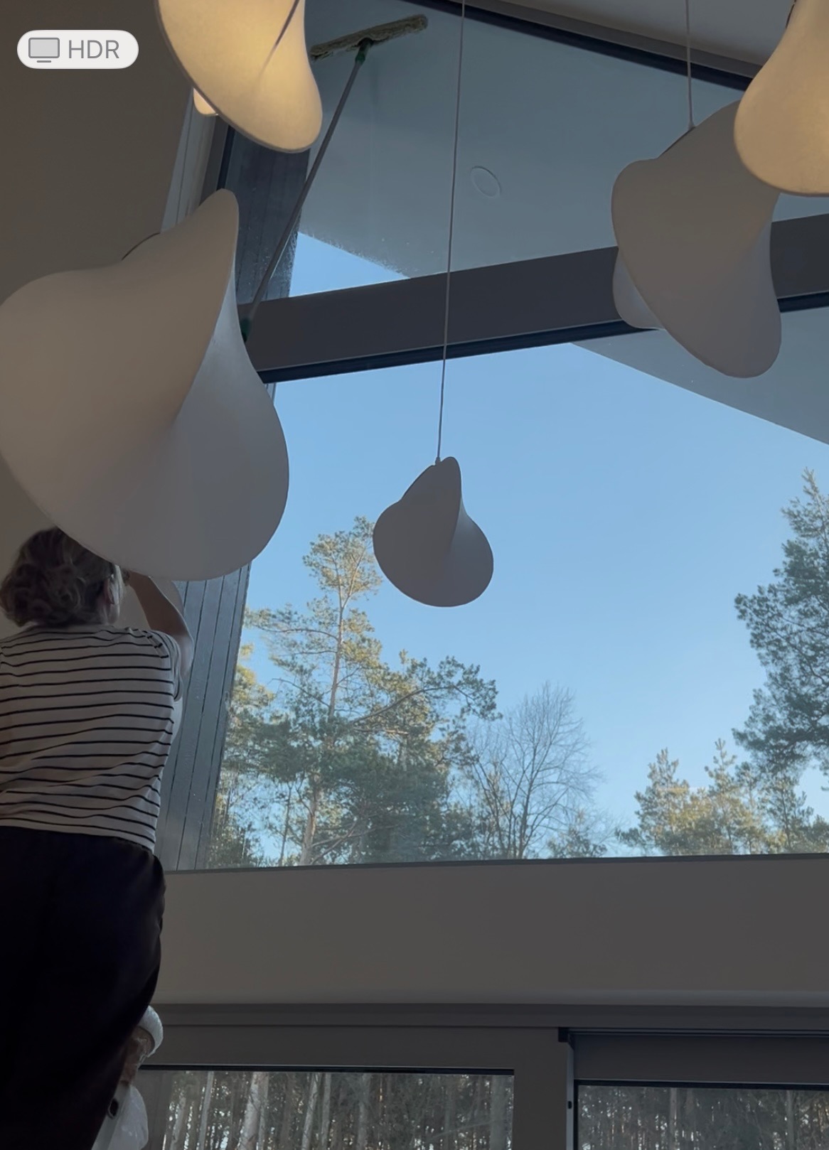 Osoba myje wysokie okno w nowoczesnym domu z widokiem na las, używając specjalistycznej myjki. Wnętrze oświetlają designerskie lampy.