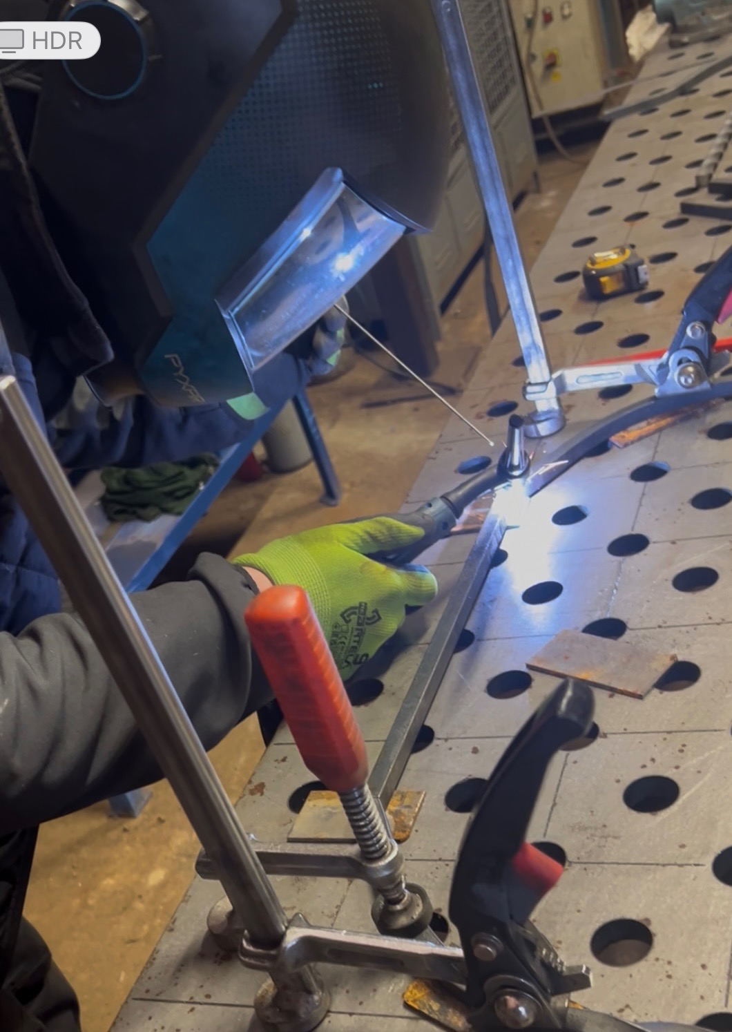 Spawanie metalowych elementów na perforowanym stole spawalniczym, widoczne iskry, uchwyty mocujące i ochronna maska spawalnicza.