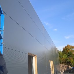 Emilian Wojtczak - Budowa hali: montaż paneli ściennych z widocznymi oknami w drewnianych ramach, niebieski podnośnik i fragment jesiennego krajobrazu w tle.