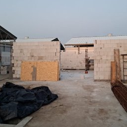 Emilian Wojtczak - Wznoszenie ścian z bloczków betonu komórkowego na placu budowy, widoczne elementy konstrukcji stalowej dachu, składowane zbrojenie i płyta OSB.