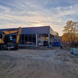 Emilian Wojtczak - Budowa nowoczesnego budynku z szkieletem stalowym w trakcie wznoszenia, z żółtą koparką JCB na pierwszym planie i pracownikiem w kamizelce odblaskowej przechodzącym obok, na tle jesiennego...