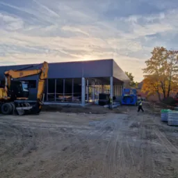 Budowa nowoczesnego budynku z szkieletem stalowym w trakcie wznoszenia, z żółtą koparką JCB na pierwszym planie i pracownikiem w kamizelce odblaskowej przechodzącym obok, na tle jesiennego...