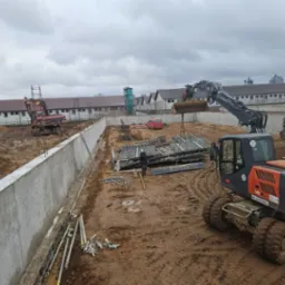 Budowa betonowego muru oporowego na placu budowy z dwoma koparkami i stosem metalowych szalunków w tle, pochmurny dzień.