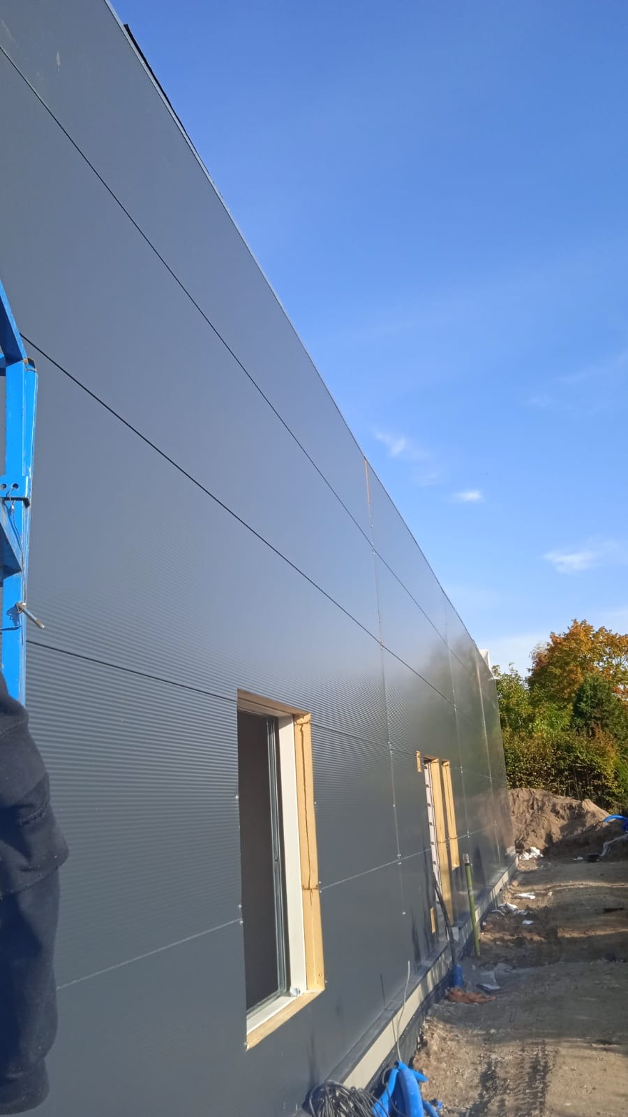 Budowa hali: montaż paneli ściennych z widocznymi oknami w drewnianych ramach, niebieski podnośnik i fragment jesiennego krajobrazu w tle.