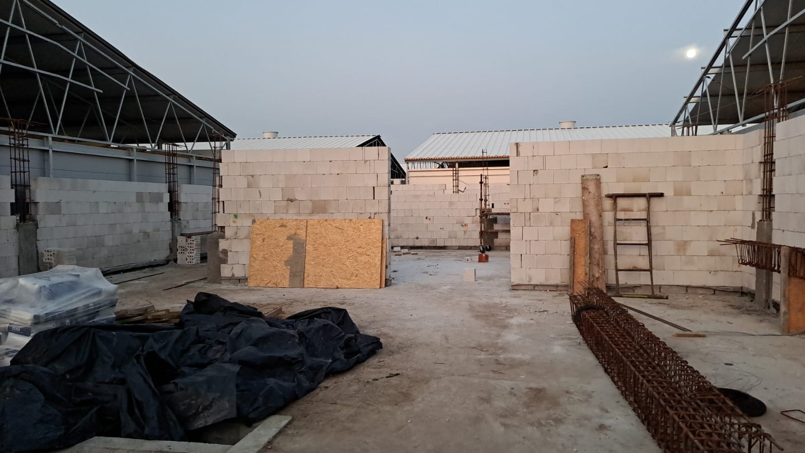 Wznoszenie ścian z bloczków betonu komórkowego na placu budowy, widoczne elementy konstrukcji stalowej dachu, składowane zbrojenie i płyta OSB.