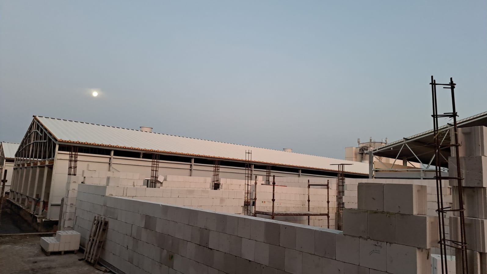 Budowa hali przemysłowej z bloczków betonu komórkowego Ytong, widoczne zbrojenia i konstrukcja dachu w trakcie montażu, księżyc na niebie.