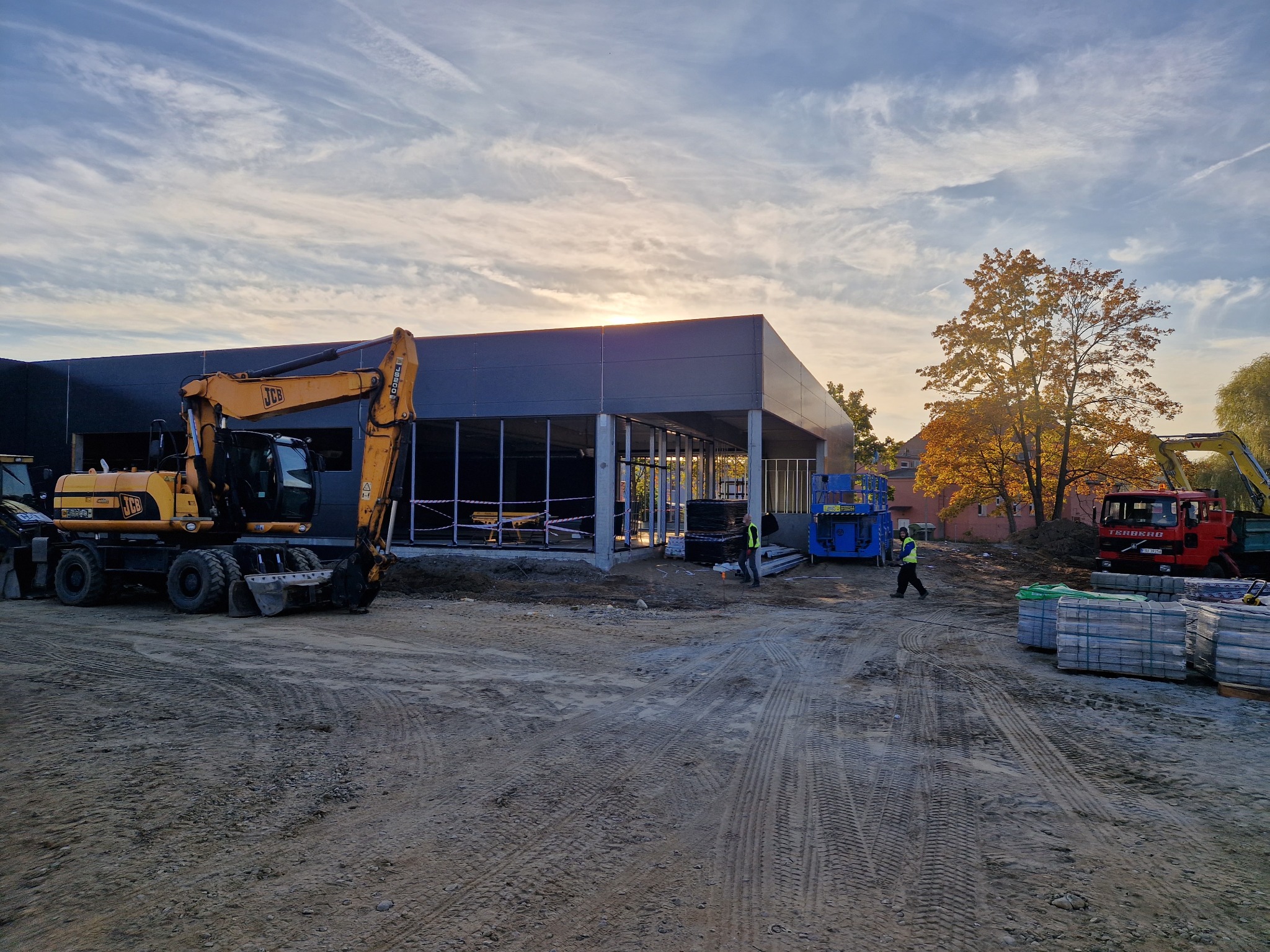 Budowa nowoczesnego budynku z szkieletem stalowym w trakcie wznoszenia, z żółtą koparką JCB na pierwszym planie i pracownikiem w kamizelce odblaskowej przechodzącym obok, na tle jesiennego...