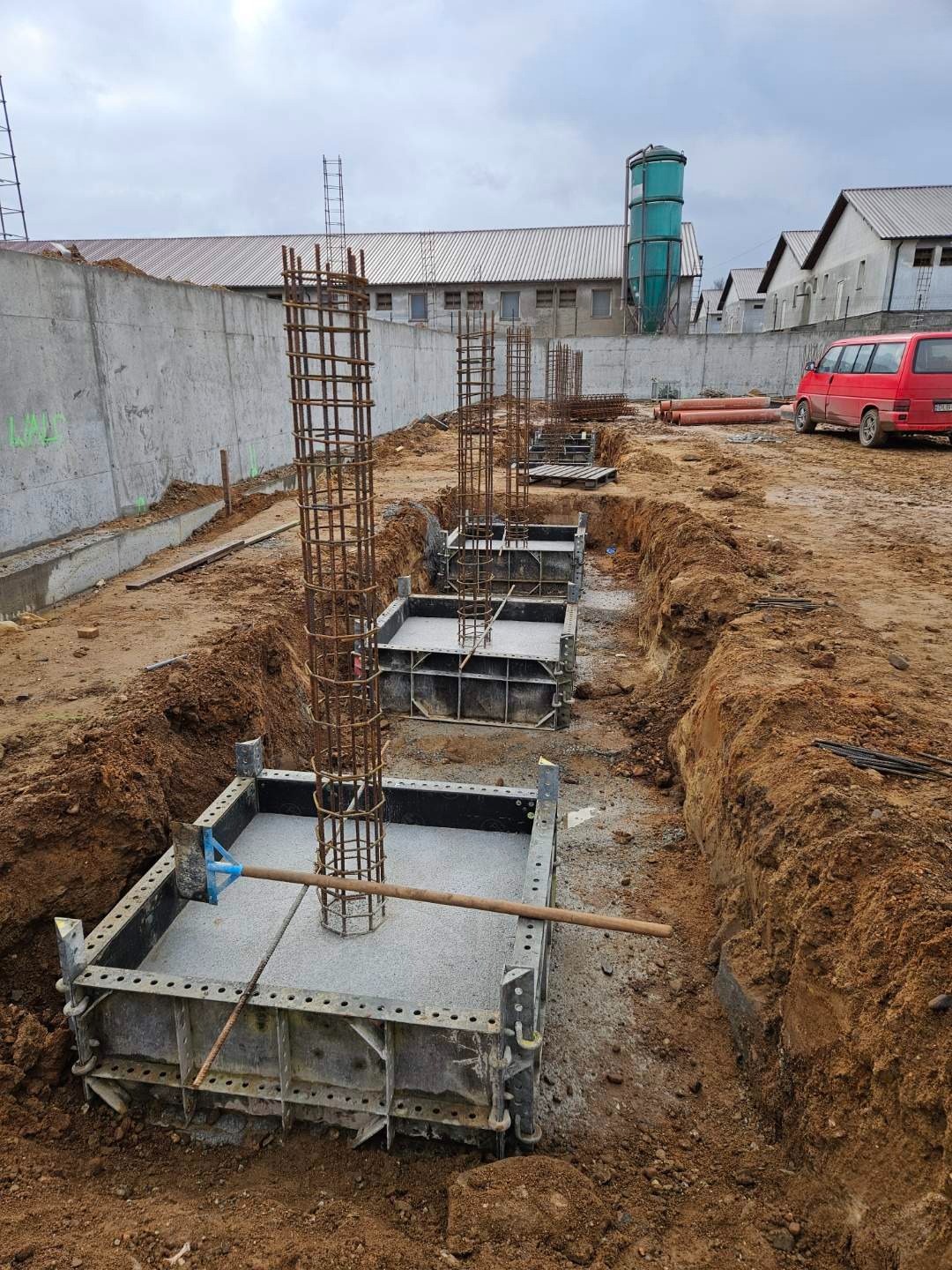 Wykopy pod fundamenty z zalewanymi betonem szalunkami i wystającymi zbrojeniami w kształcie klatek, widoczny czerwony samochód w tle.