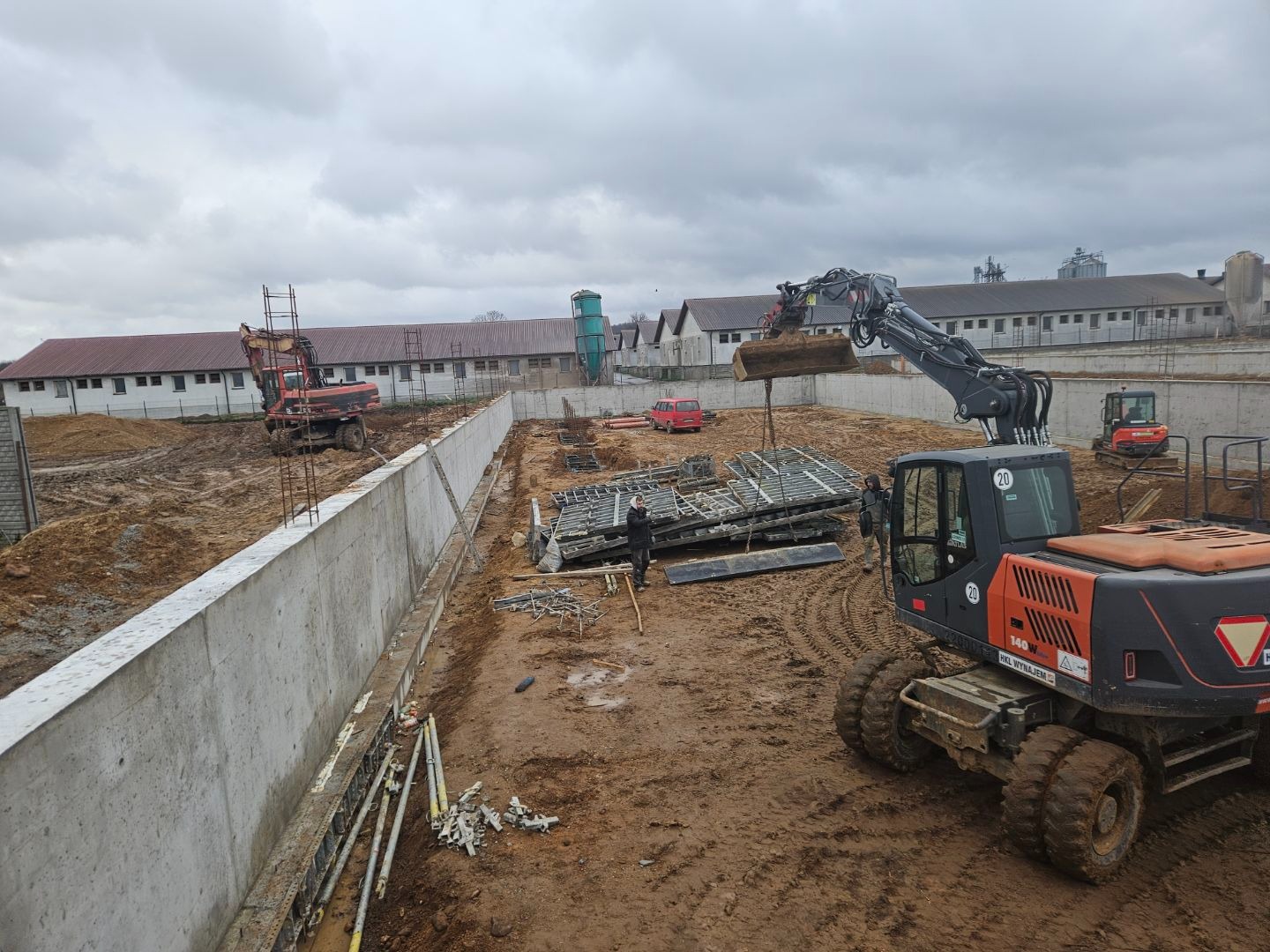 Budowa betonowego muru oporowego na placu budowy z dwoma koparkami i stosem metalowych szalunków w tle, pochmurny dzień.