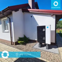 Biała pompa ciepła powietrze-woda zamontowana na betonowych podstawach przy ścianie domu z jasną elewacją i brązowym dachem, otoczona kamieniami ozdobnymi.