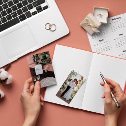 Konsultantka Ślubna Marta Wółkiewicz - Planowanie ślubu: otwarty notatnik, kalendarz z zaznaczonymi miesiącami, zdjęcia pary młodej i dekoracji stołu weselnego, pierścionek zaręczynowy, laptop, bawełna i pióro na różowym tle.