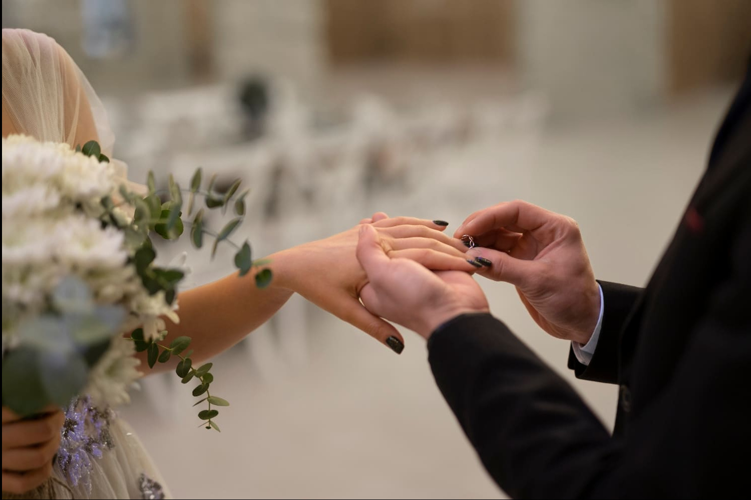 Moment zaręczyn: dłoń mężczyzny w garniturze wkłada pierścionek na palec kobiety trzymającej bukiet białych kwiatów, manicure w ciemnym kolorze.