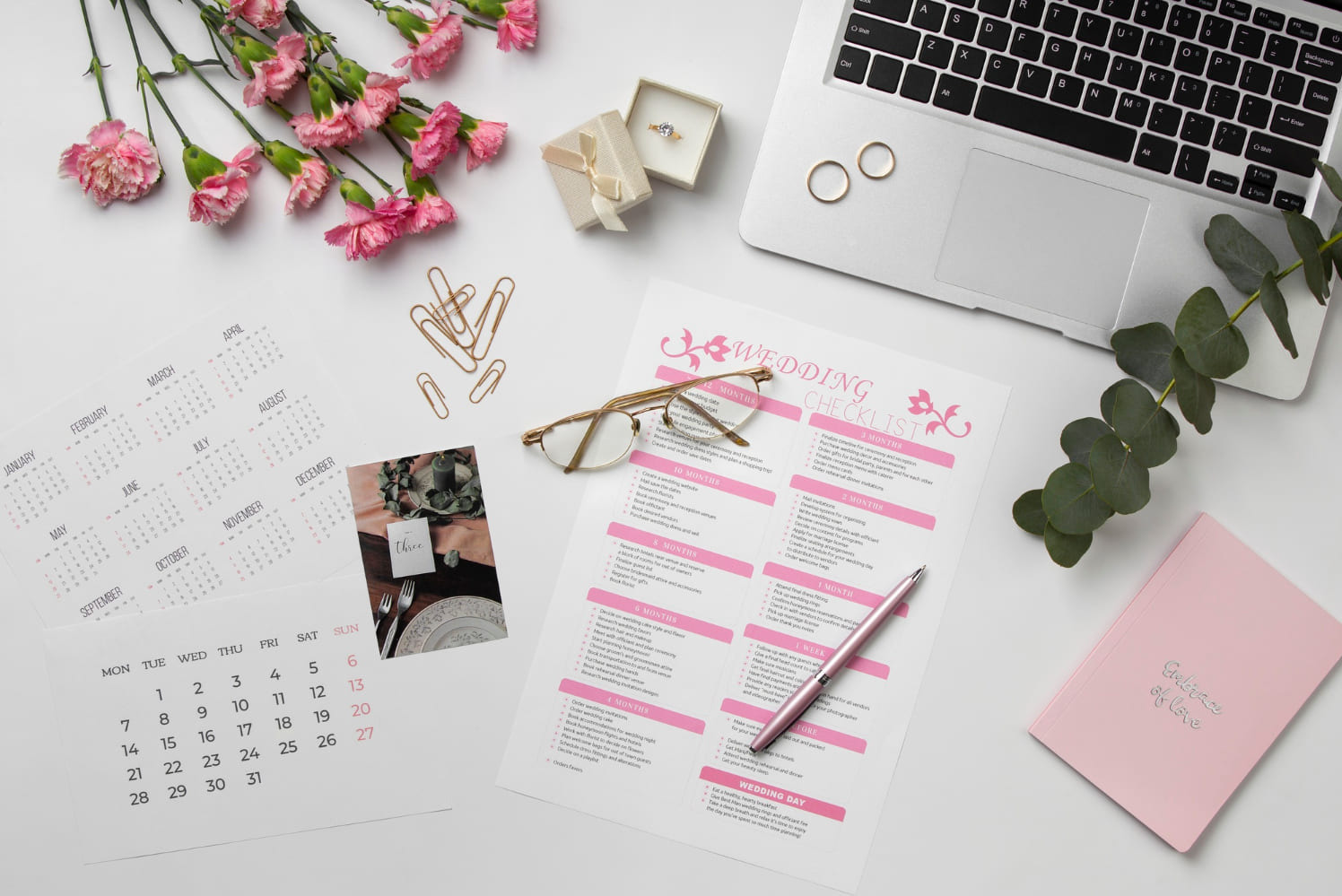 Planowanie ślubu: checklist z kalendarzem, obrączki, pierścionek zaręczynowy, notatnik z napisem 'Embrace of love', laptop i kwiaty na białym tle.
