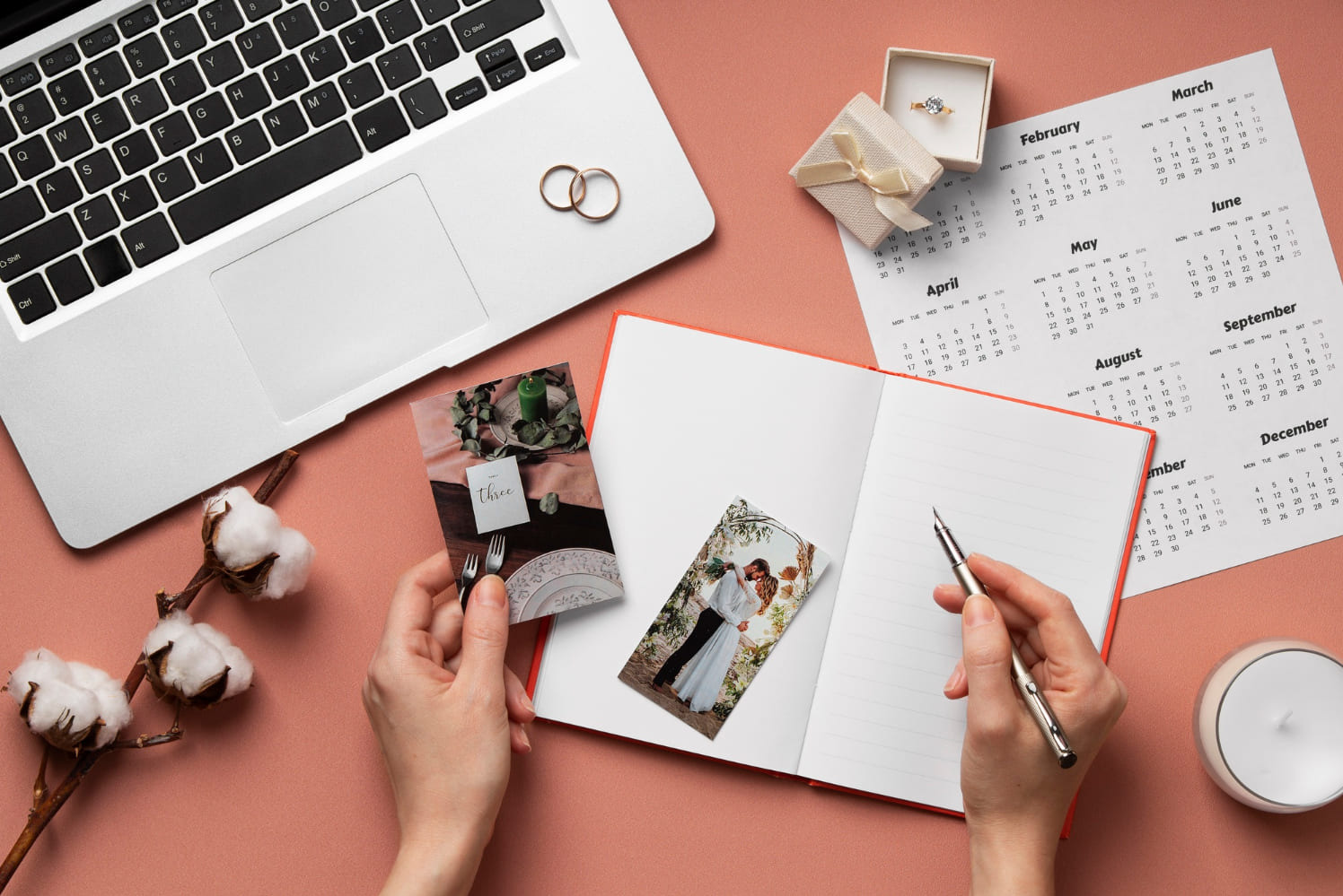 Planowanie ślubu: otwarty notatnik, kalendarz z zaznaczonymi miesiącami, zdjęcia pary młodej i dekoracji stołu weselnego, pierścionek zaręczynowy, laptop, bawełna i pióro na różowym tle.