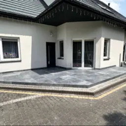 Wyłożony szarymi płytami taras przed domem z białymi ścianami i czarnym dachem, widok z poziomu kostki brukowej.