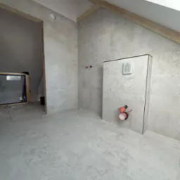 Szara łazienka na poddaszu w trakcie wykańczania, widoczne ułożone płytki podłogowe i ścienne, zabudowa stelaża WC z zamontowaną instalacją hydrauliczną oraz okno dachowe.