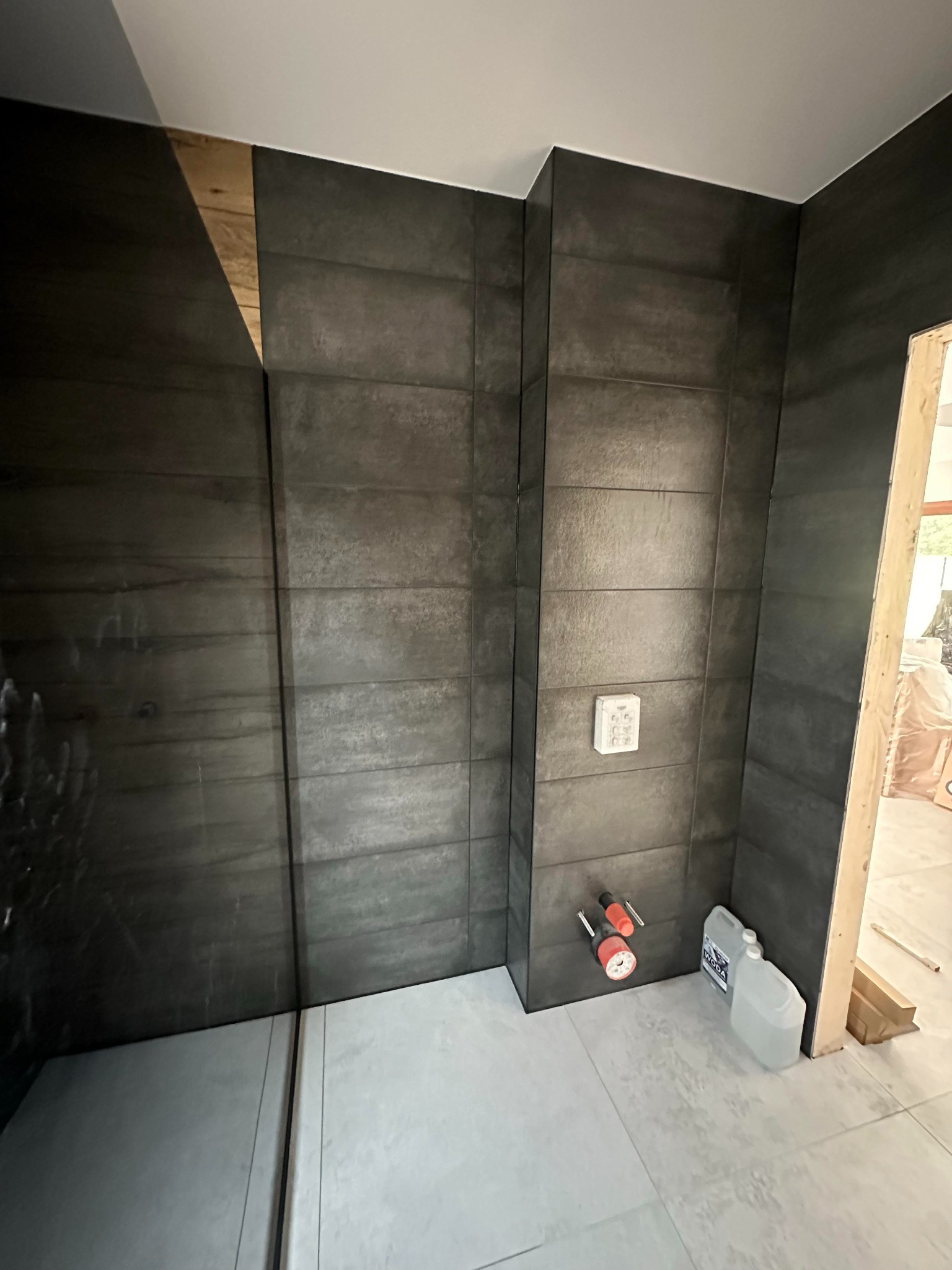 Ciemnoszare, poziome płytki imitujące kamień na ścianie kabiny prysznicowej, z widocznymi przyłączami wodnymi i puszką elektryczną, biała podłoga i częściowo widoczne drewniane elementy konstrukcyjne
