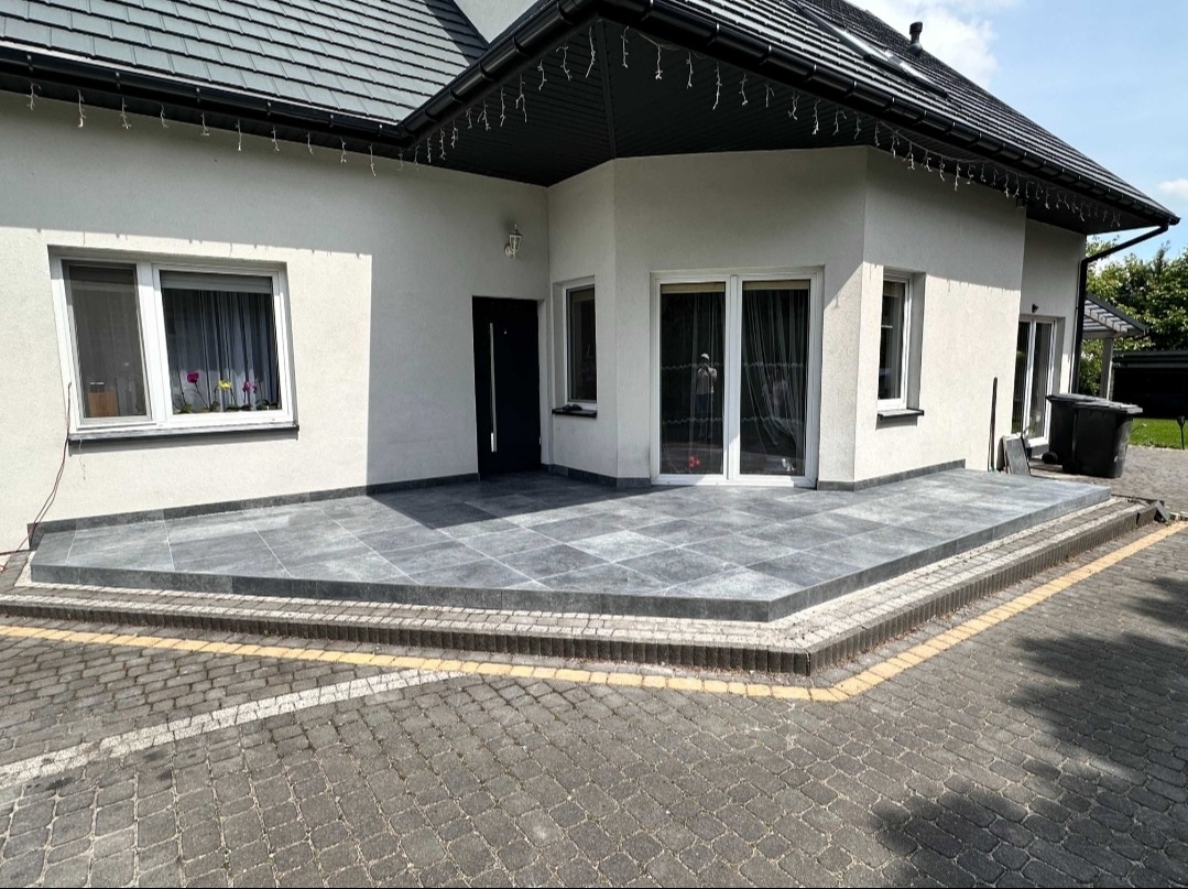 Wyłożony szarymi płytami taras przed domem z białymi ścianami i czarnym dachem, widok z poziomu kostki brukowej.