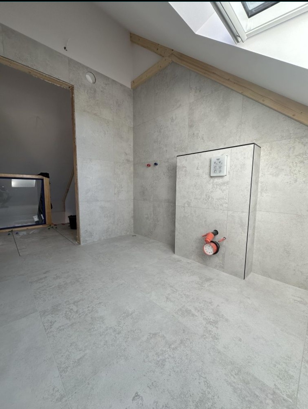 Szara łazienka na poddaszu w trakcie wykańczania, widoczne ułożone płytki podłogowe i ścienne, zabudowa stelaża WC z zamontowaną instalacją hydrauliczną oraz okno dachowe.