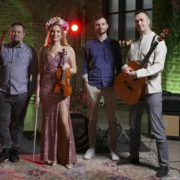 Zespół muzyczny w składzie: basista z basem, skrzypaczka w kwiecistym wianku trzymająca skrzypce, gitarzysta z gitarą akustyczną oraz perkusista w tle, na tle ceglanej ściany z zielonym oświetleniem.