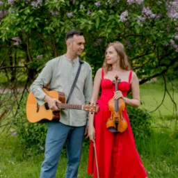 Duet muzyczny: mężczyzna z gitarą akustyczną i kobieta w czerwonej sukience z skrzypcami, na tle kwitnącego bzu.