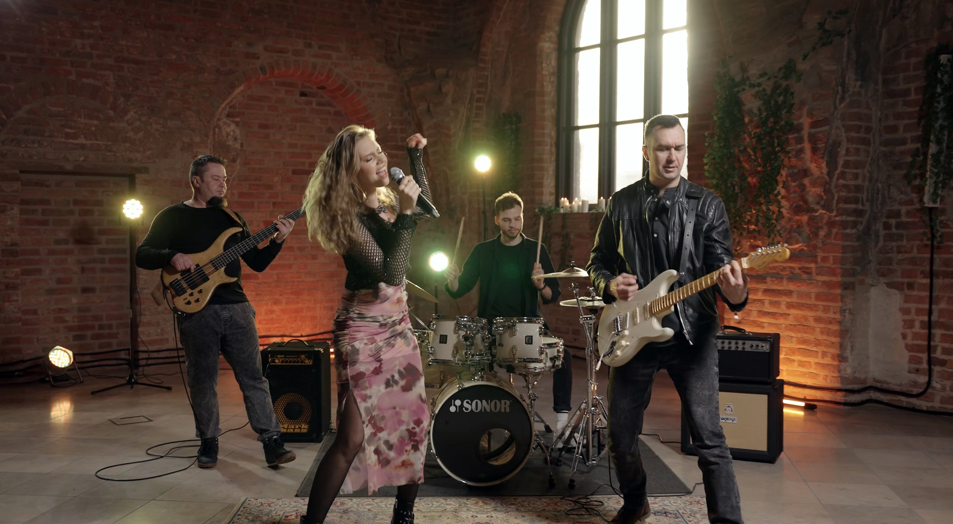 Zespół muzyczny w trakcie występu w ceglanym wnętrzu, wokalistka z mikrofonem, gitarzysta w skórzanej kurtce, basista i perkusista, oświetlenie sceniczne.