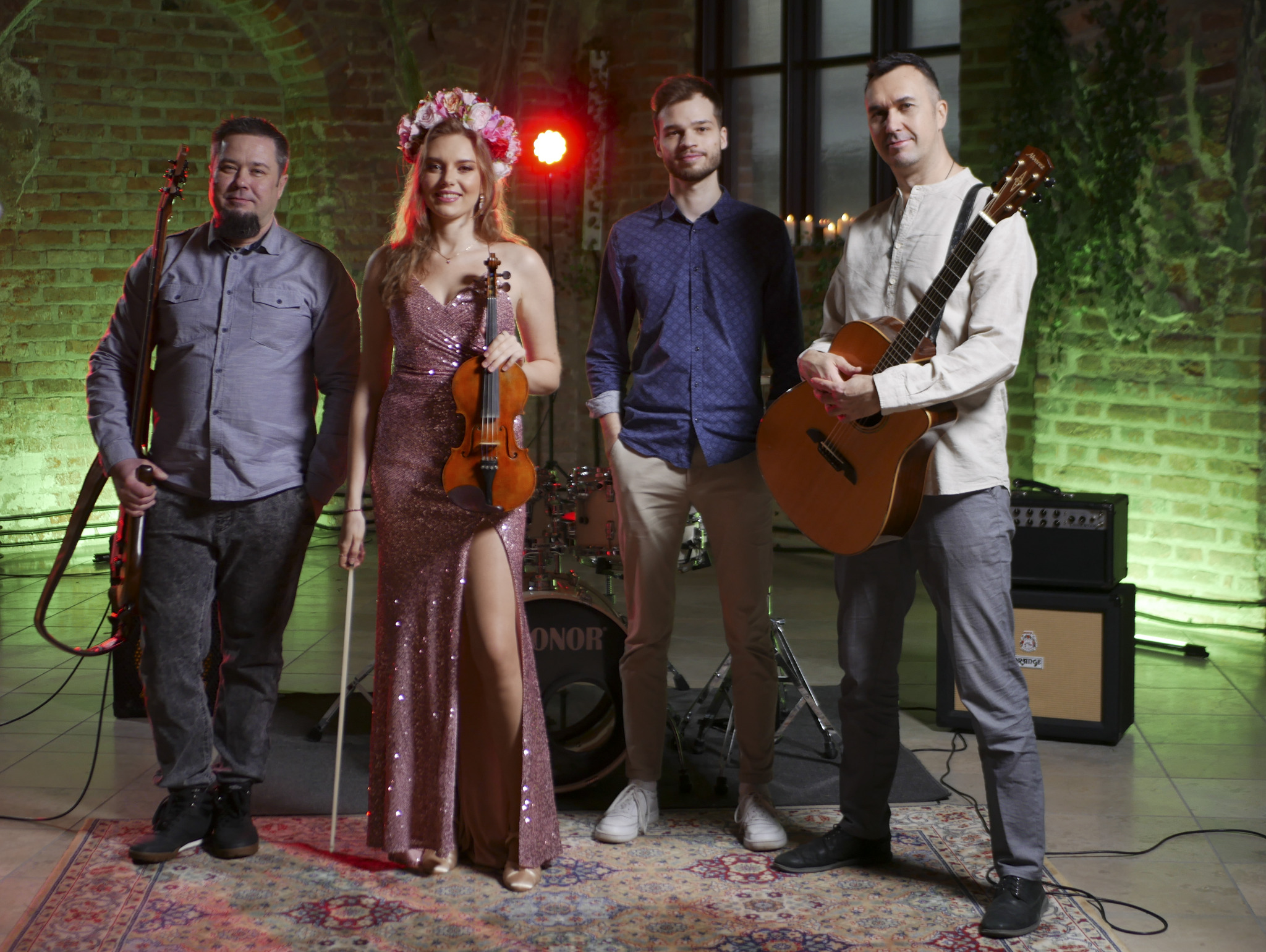 Zespół muzyczny w składzie: basista z basem, skrzypaczka w kwiecistym wianku trzymająca skrzypce, gitarzysta z gitarą akustyczną oraz perkusista w tle, na tle ceglanej ściany z zielonym oświetleniem.