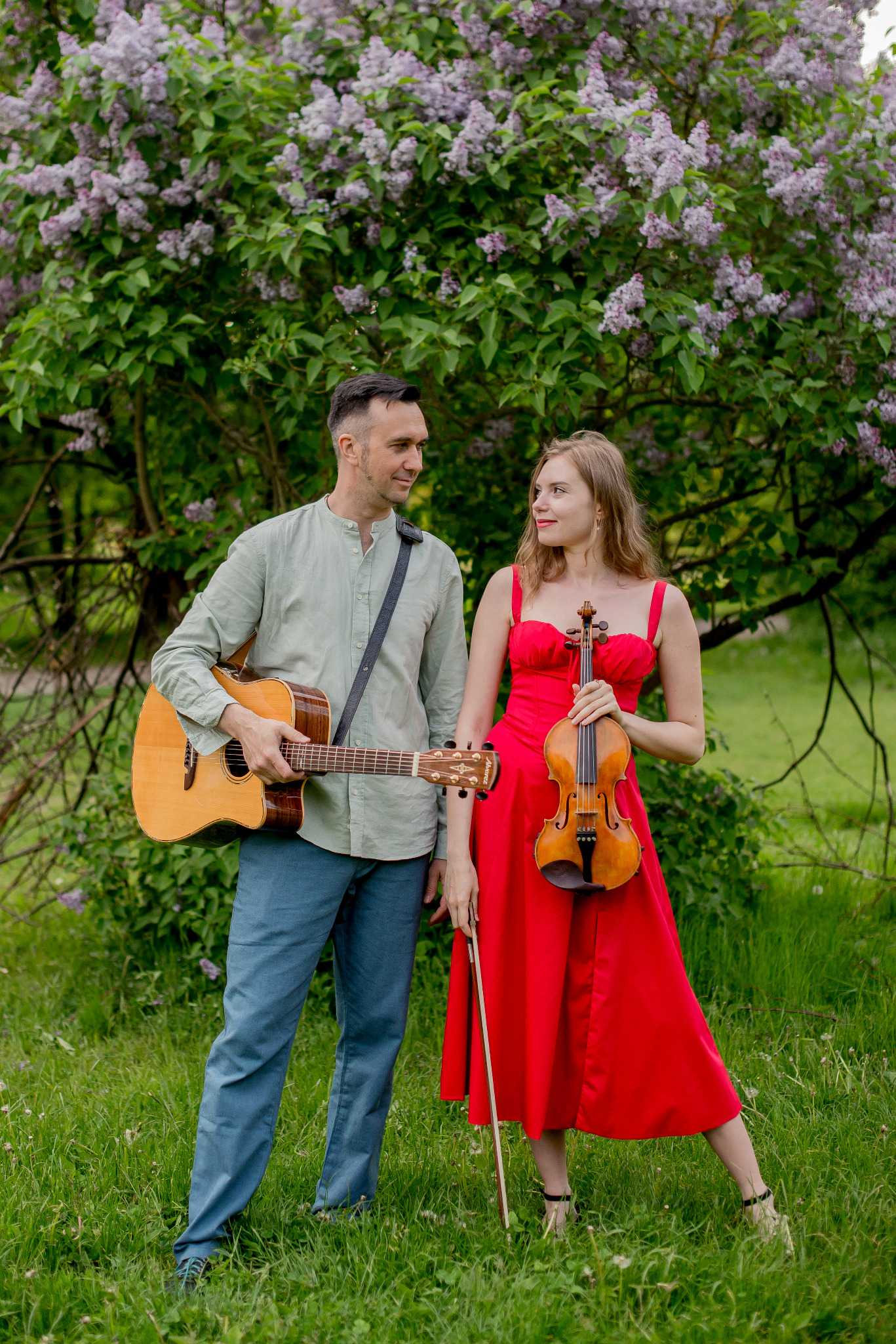 Duet muzyczny: mężczyzna z gitarą akustyczną i kobieta w czerwonej sukience z skrzypcami, na tle kwitnącego bzu.