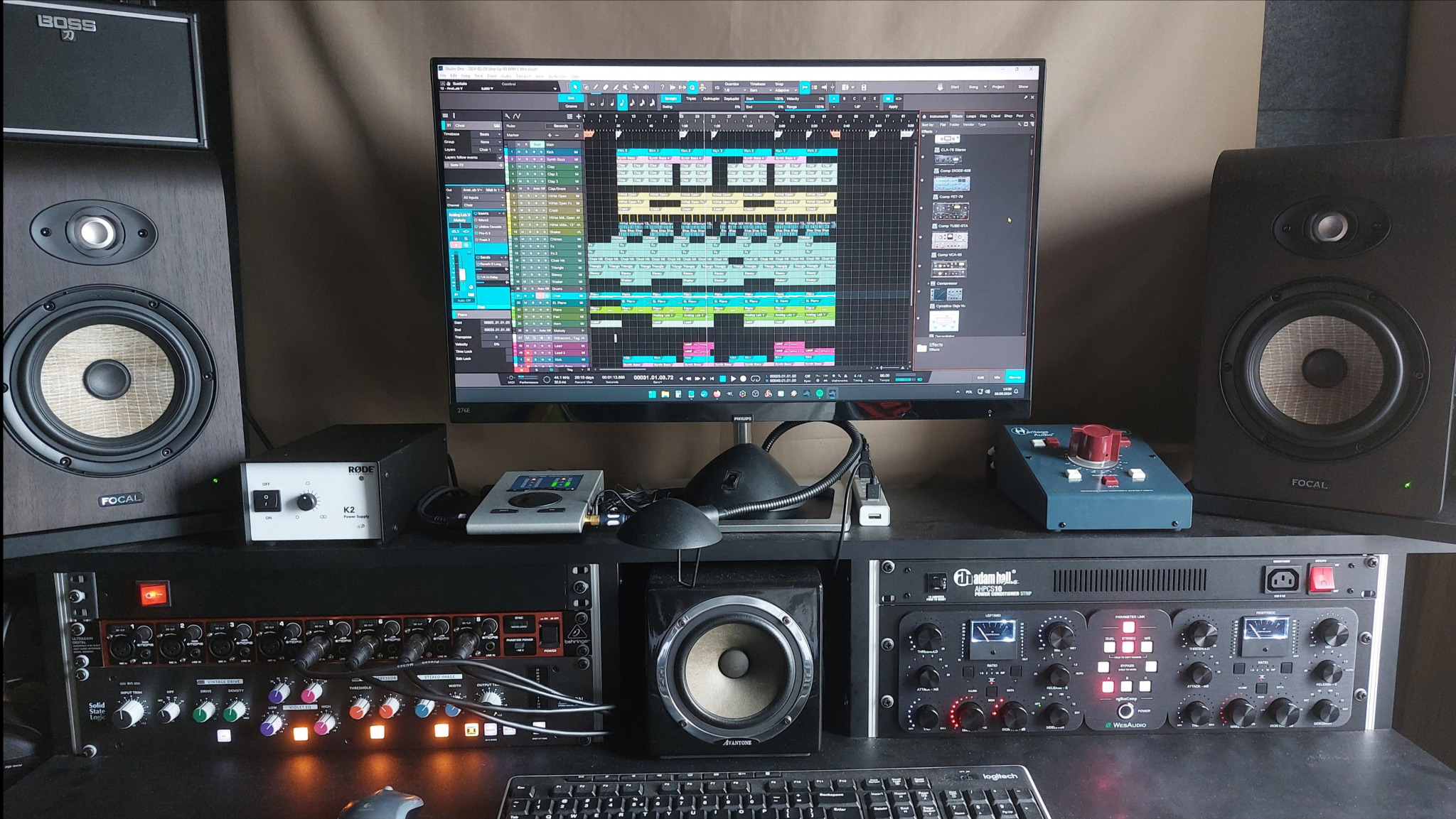 Widok na monitor z oprogramowaniem DAW prezentującym układ ścieżek audio, otoczony przez monitory studyjne Focal i Boss, procesory dźwięku SSL i Adam Hall, interfejs audio RODE K2 oraz mikrofon...