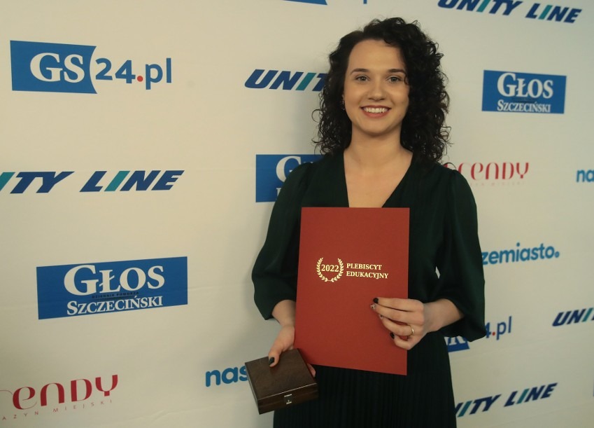 Uśmiechnięta kobieta w zielonej sukience trzyma dyplom 'Plebiscyt Edukacyjny 2022' i małe drewniane pudełko na tle ściany z logotypami.