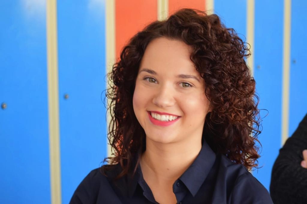 Portret uśmiechniętej kobiety z brązowymi kręconymi włosami i czerwonymi ustami na tle niebiesko-pomarańczowych paneli.