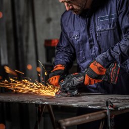DNIPRO-M STORES SPÓŁKA Z OGRANICZONĄ ODPOWIEDZIALNOŚCIĄ - Spawacz w kombinezonie i rękawicach ochronnych szlifuje metalową płytę szlifierką kątową, iskry rozbryzgują się na boki.