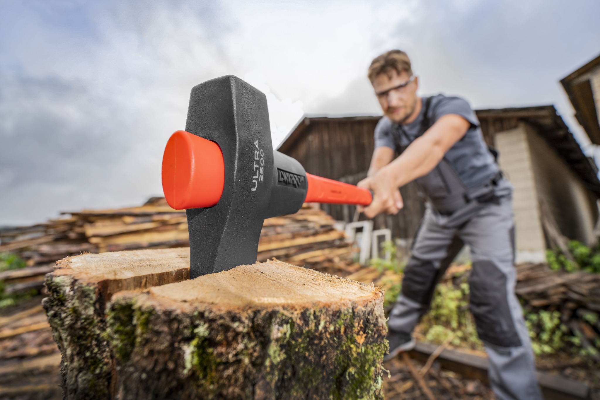 Mężczyzna w okularach ochronnych zamachuje się toporem z pomarańczowym trzonkiem na pniu, w tle sterta drewna i budynek gospodarczy.