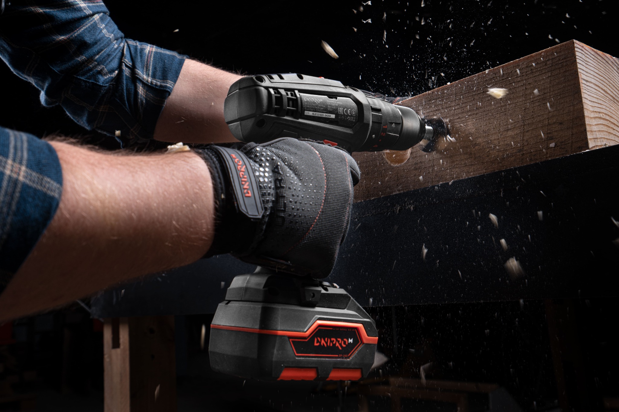 Mężczyzna w rękawicach ochronnych wierci otwór w drewnianej belce za pomocą wiertarki akumulatorowej, wióry w powietrzu.