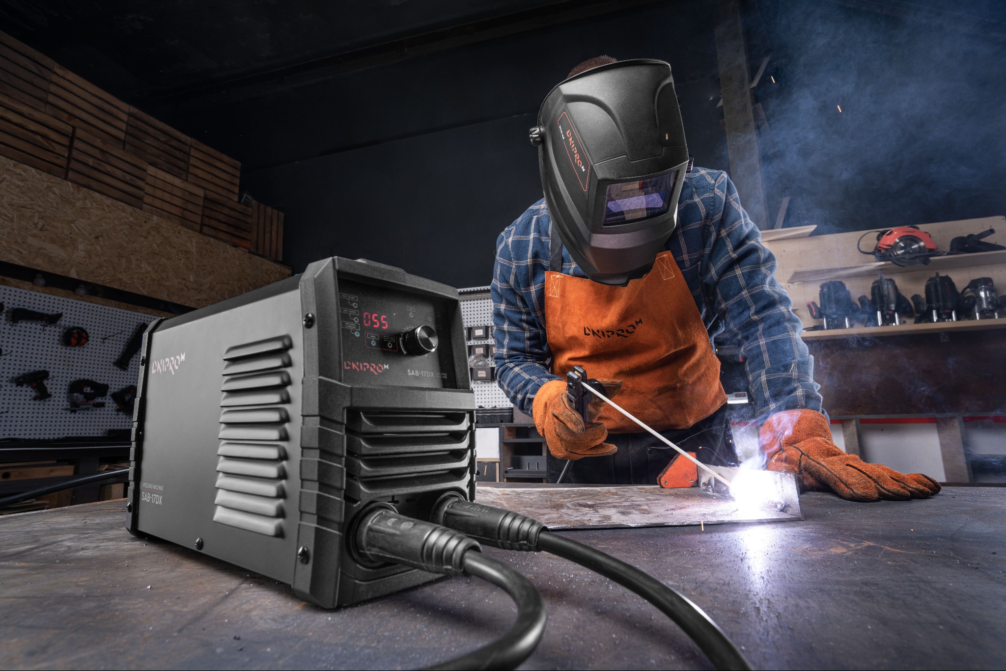 Spawacz w masce ochronnej i skórzanym fartuchu, spawający metal na warsztatowym stole obok spawarki UNIPROM SAB-17DX, w tle narzędzia na ścianie.