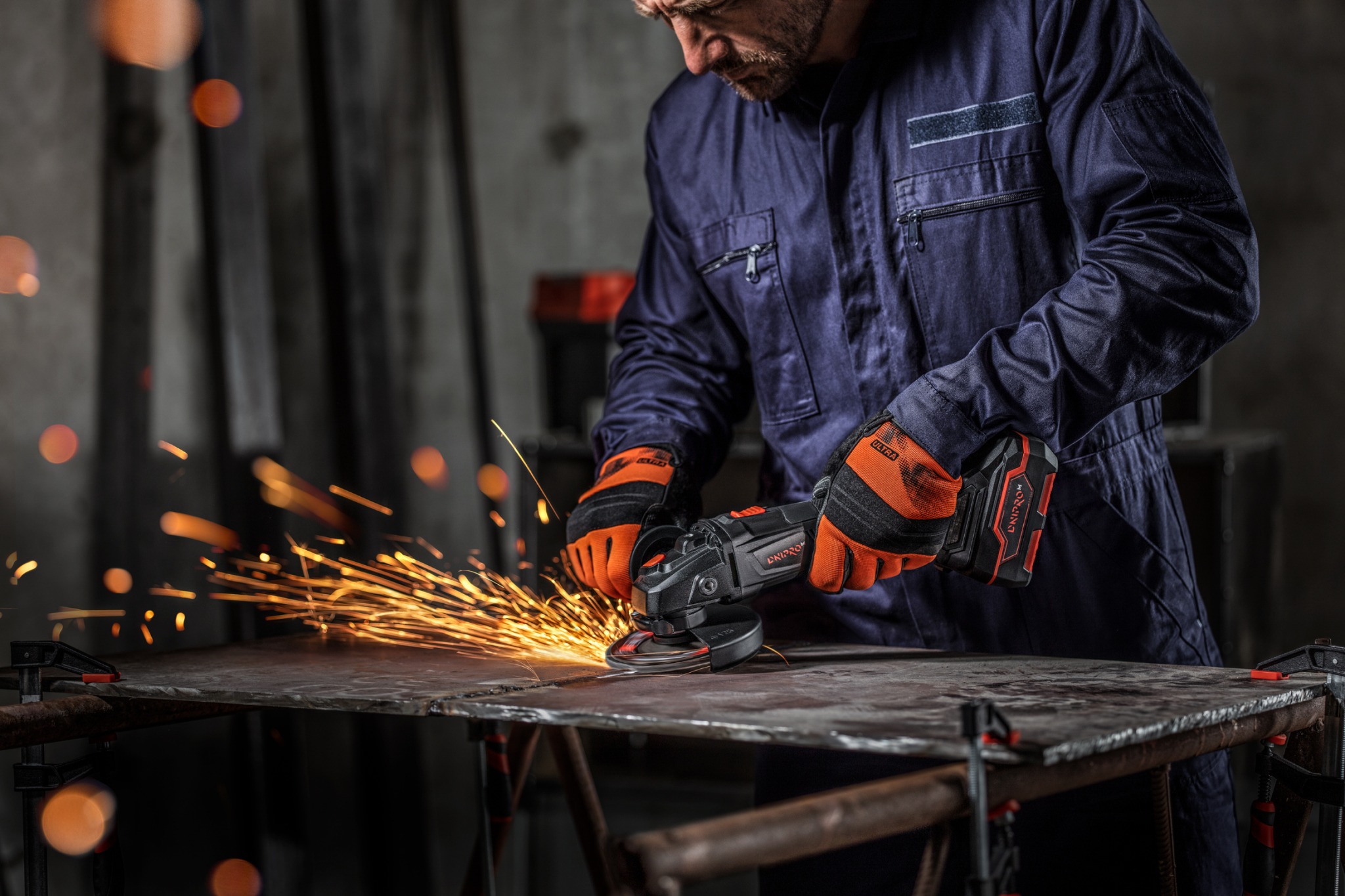 Spawacz w kombinezonie i rękawicach ochronnych szlifuje metalową płytę szlifierką kątową, iskry rozbryzgują się na boki.