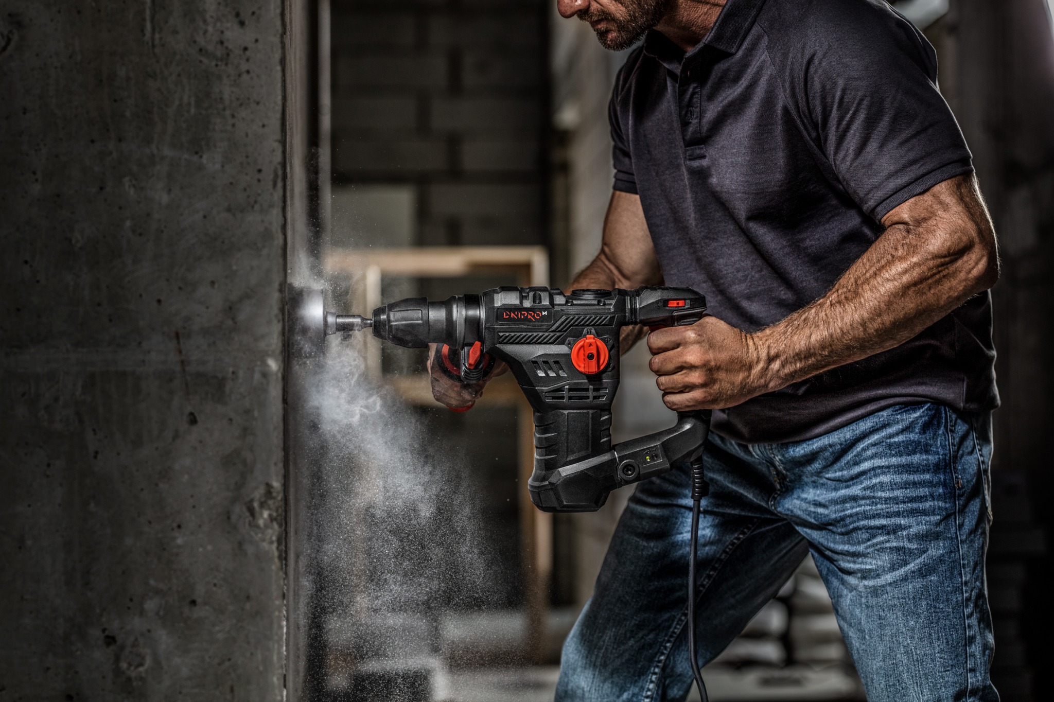 Mężczyzna wierci otwór w betonowej ścianie wiertarką udarową marki Dnipro-M, unoszący się pył, widoczne zarysowania na ścianie.