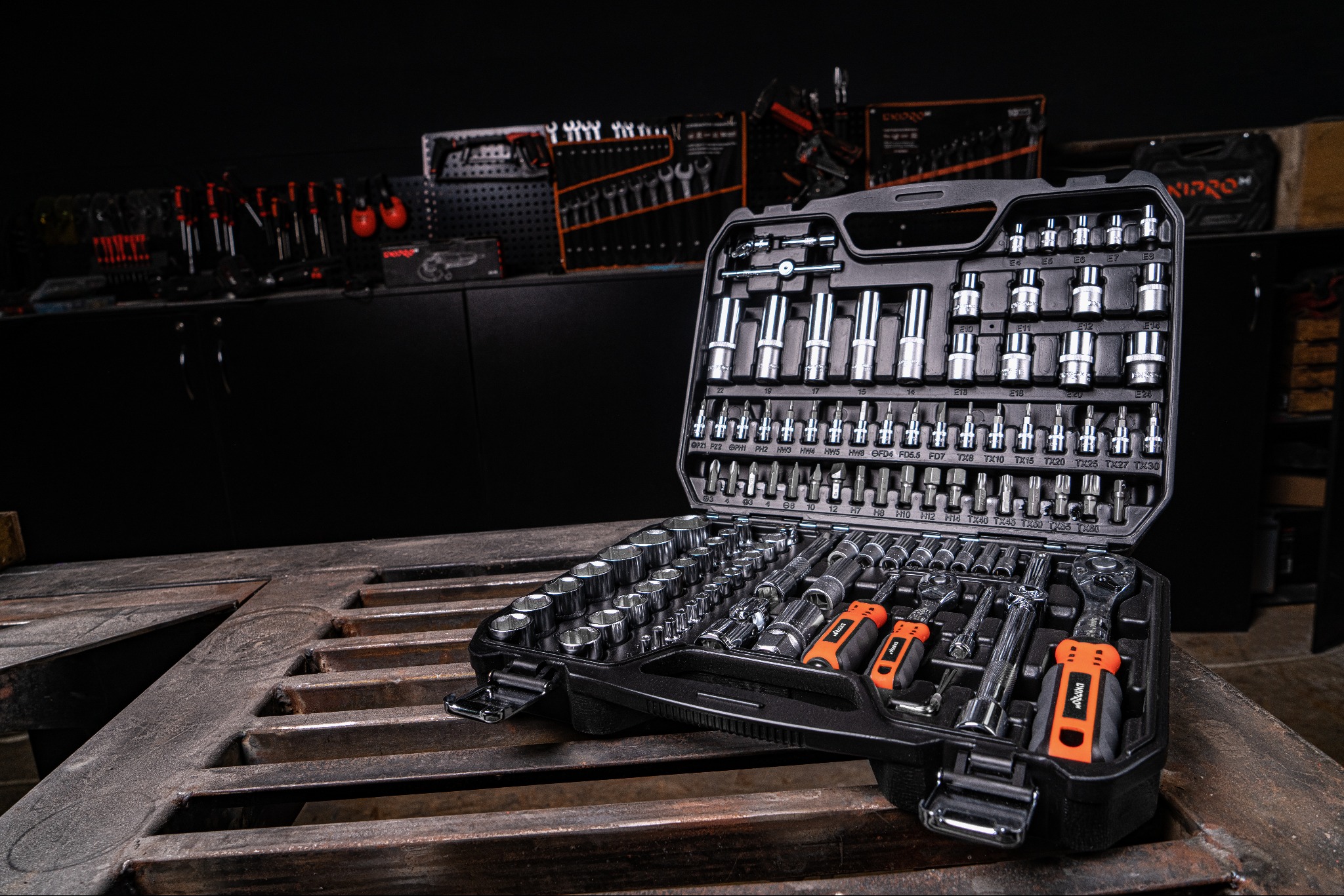 Otwarty czarny zestaw narzędzi z chromowanymi nasadkami i pomarańczowymi uchwytami na metalowym perforowanym stole warsztatowym, narzędzia wiszą na ścianie w tle.