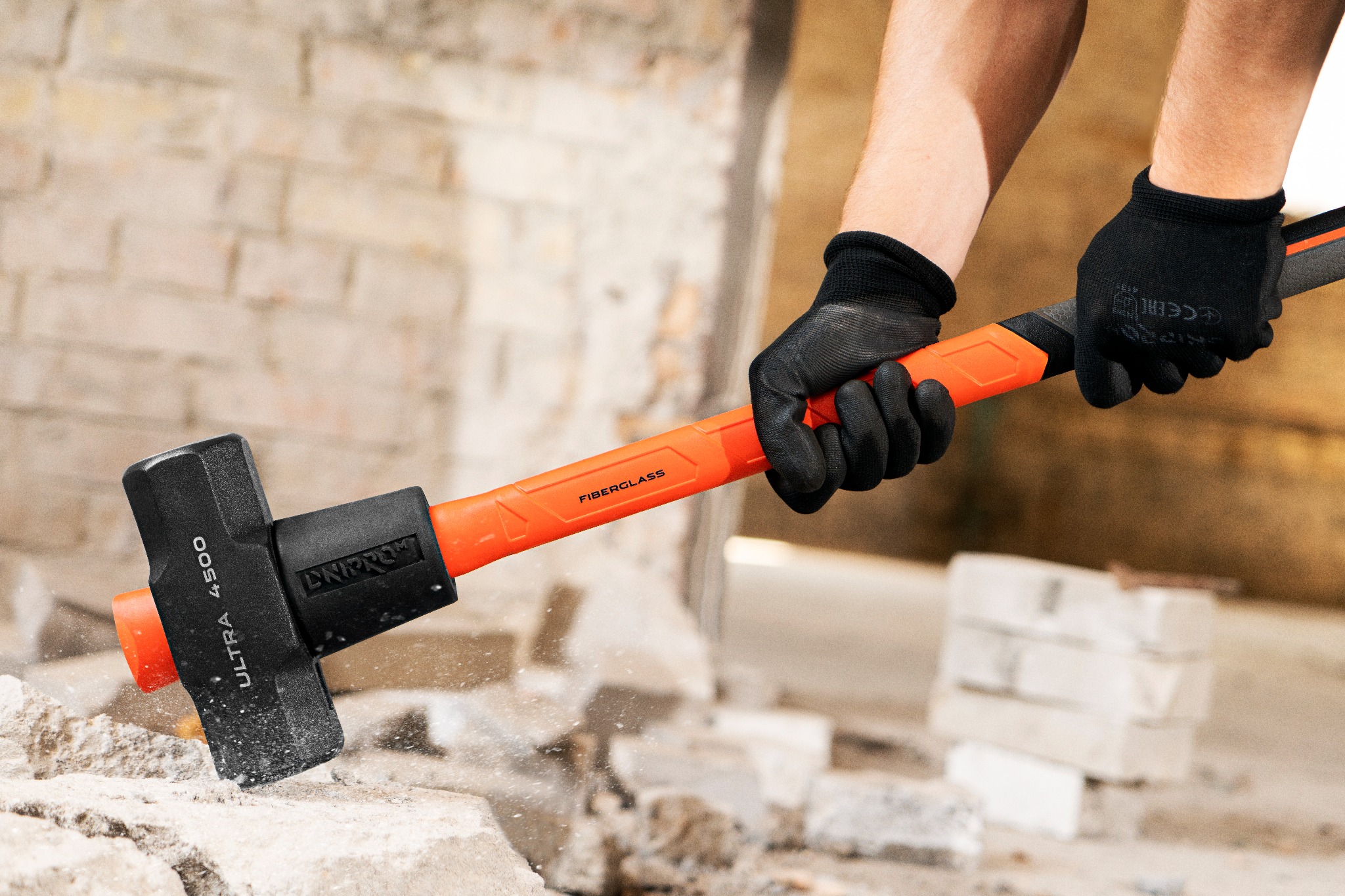 Mężczyzna w czarnych rękawicach uderza młotem o pomarańczowej rękojeści w gruz ceglany, w tle ściana z cegieł.