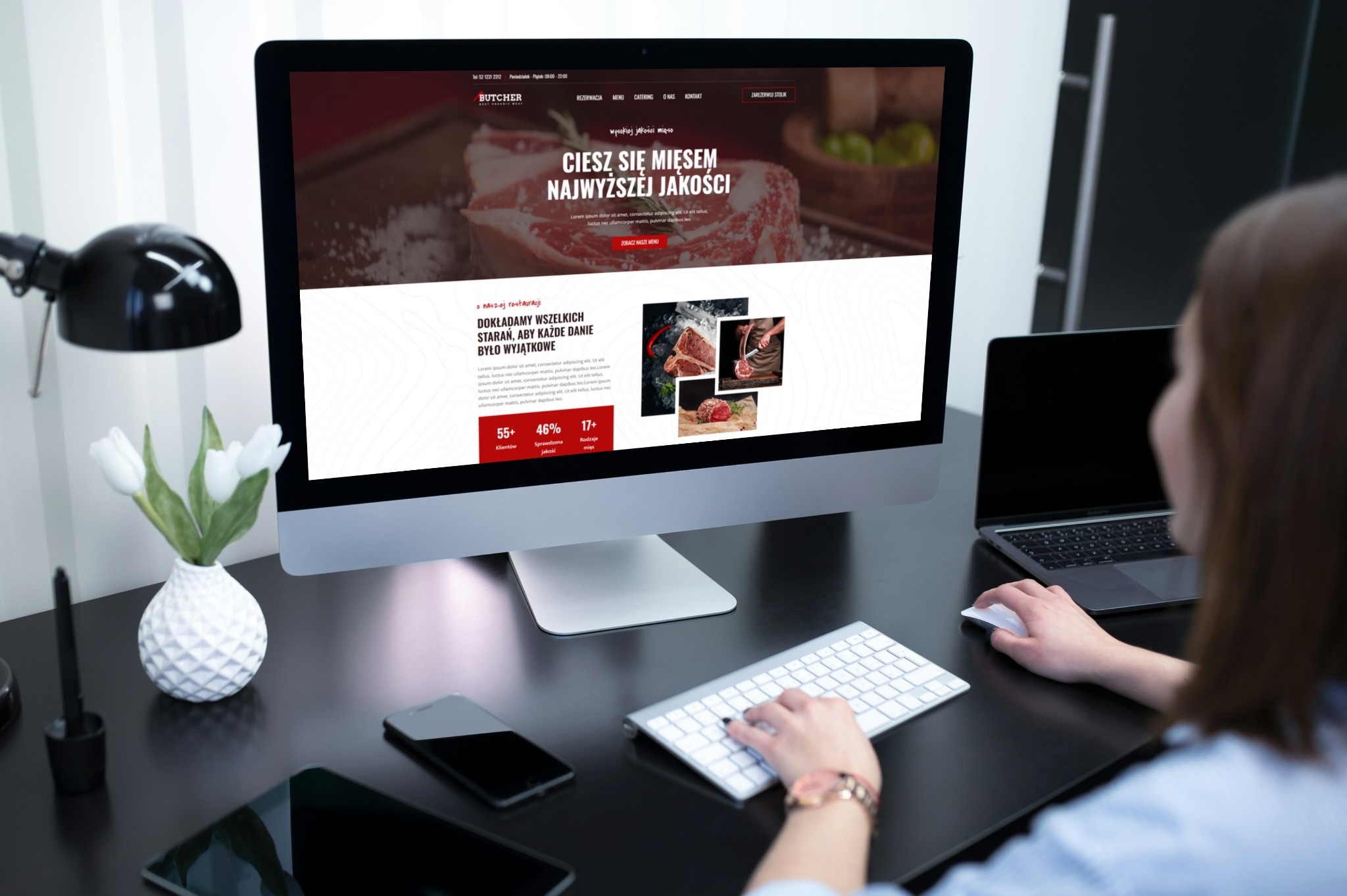 Kobieta pracuje przy biurku z komputerem wyświetlającym stronę internetową restauracji mięsnej. Widoczne tulipany, smartfon i laptop.