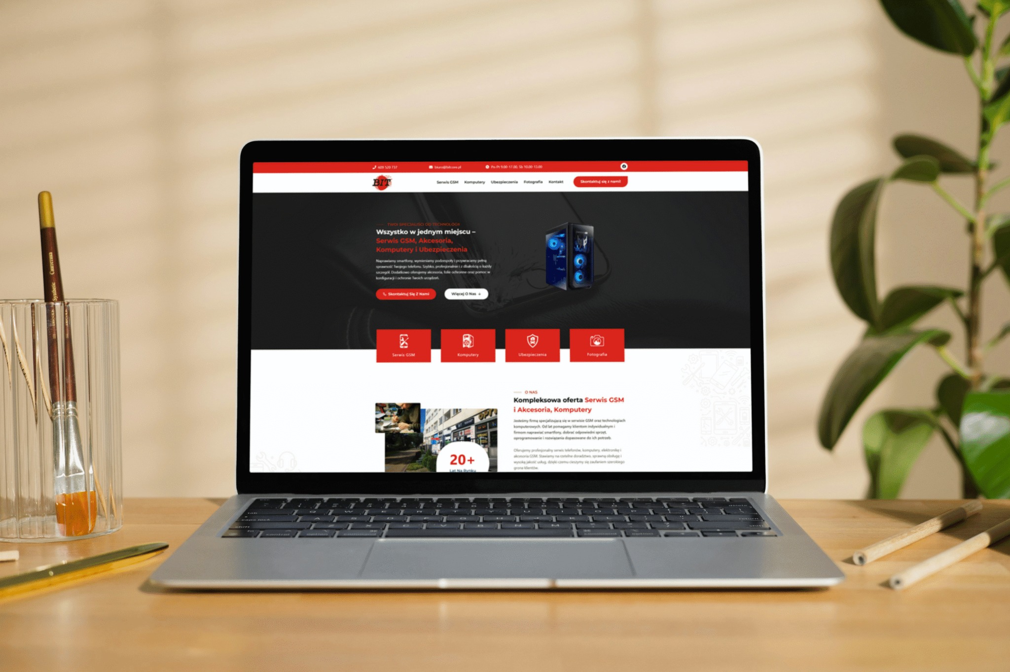 Laptop prezentuje stronę internetową firmy BIT z ofertą serwisu GSM, komputerów i ubezpieczeń. Na biurku pędzle w szklance i ołówki. W tle roślina doniczkowa.