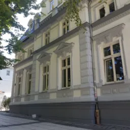 Odnowiona elewacja kamienicy z szarymi elementami dekoracyjnymi, widoczny fragment chodnika i drzewa