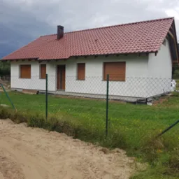 Nowo wybudowany dom jednorodzinny z czerwoną dachówką, białymi ścianami i brązowymi roletami, widziany zza zielonego ogrodzenia z siatki.