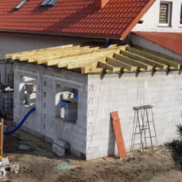 Wznoszenie ścian budynku z bloczków betonowych z widoczną konstrukcją drewnianą dachu, rury wentylacyjne i drabina oparta o ścianę.