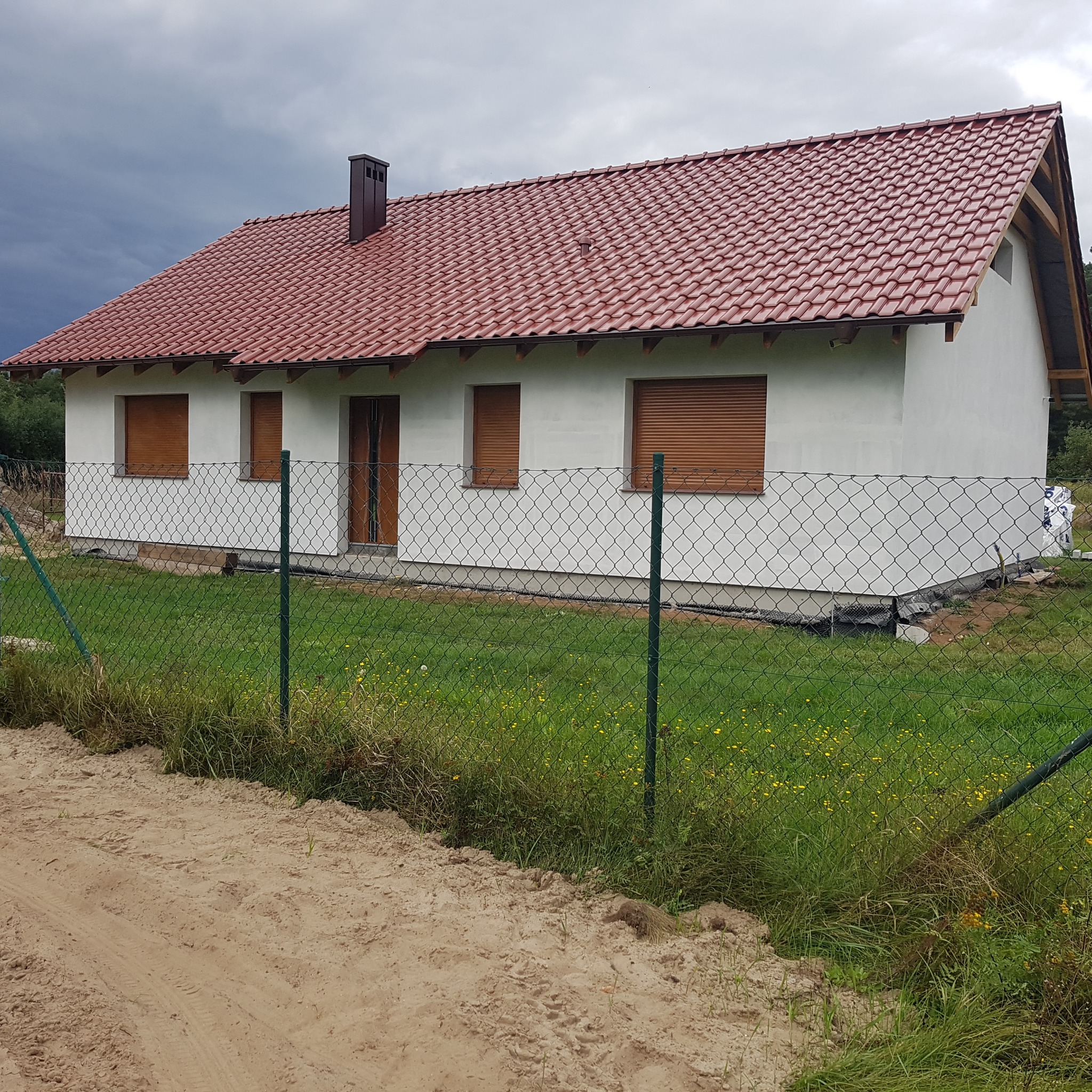 Nowo wybudowany dom jednorodzinny z czerwoną dachówką, białymi ścianami i brązowymi roletami, widziany zza zielonego ogrodzenia z siatki.