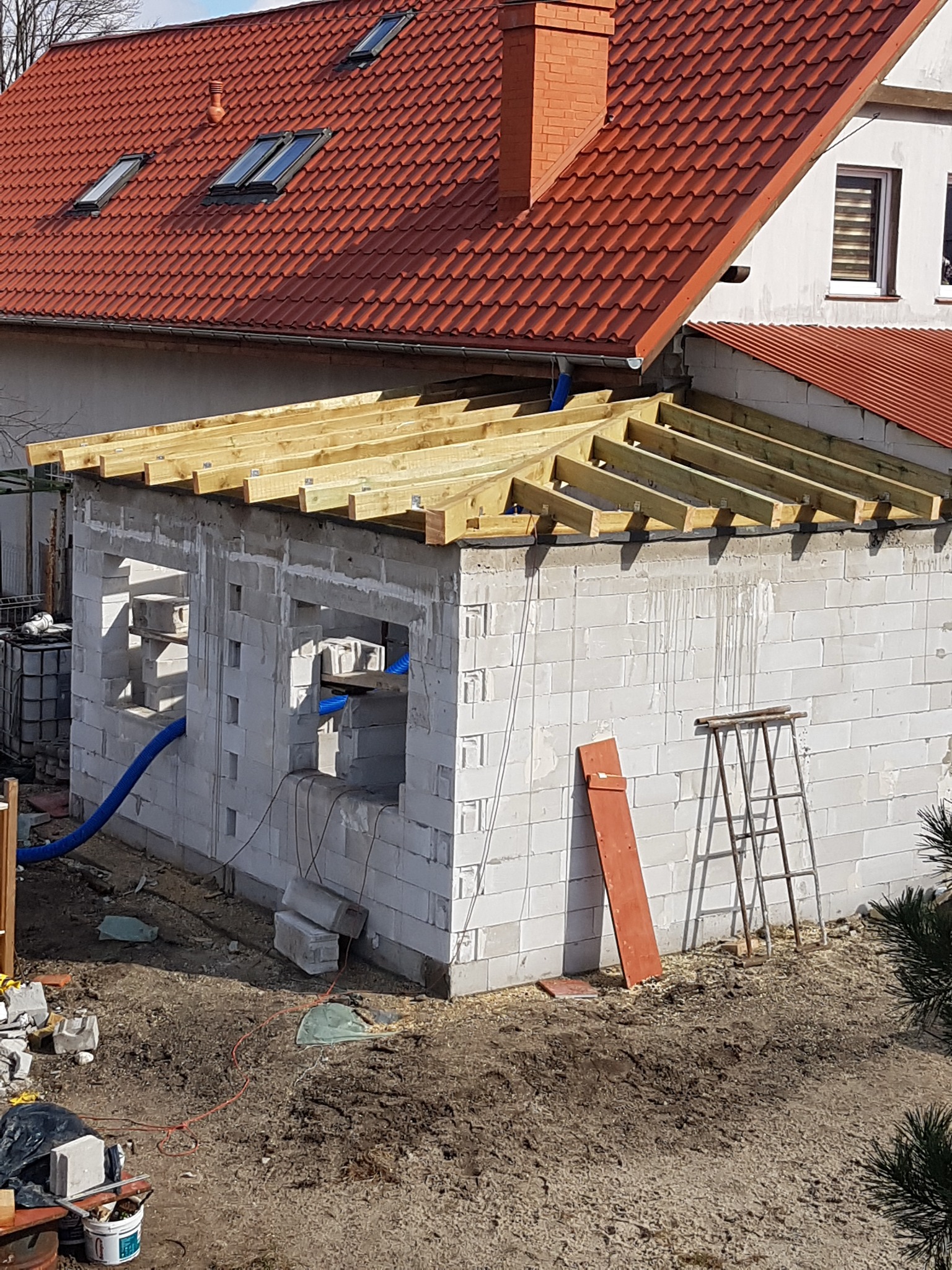 Wznoszenie ścian budynku z bloczków betonowych z widoczną konstrukcją drewnianą dachu, rury wentylacyjne i drabina oparta o ścianę.