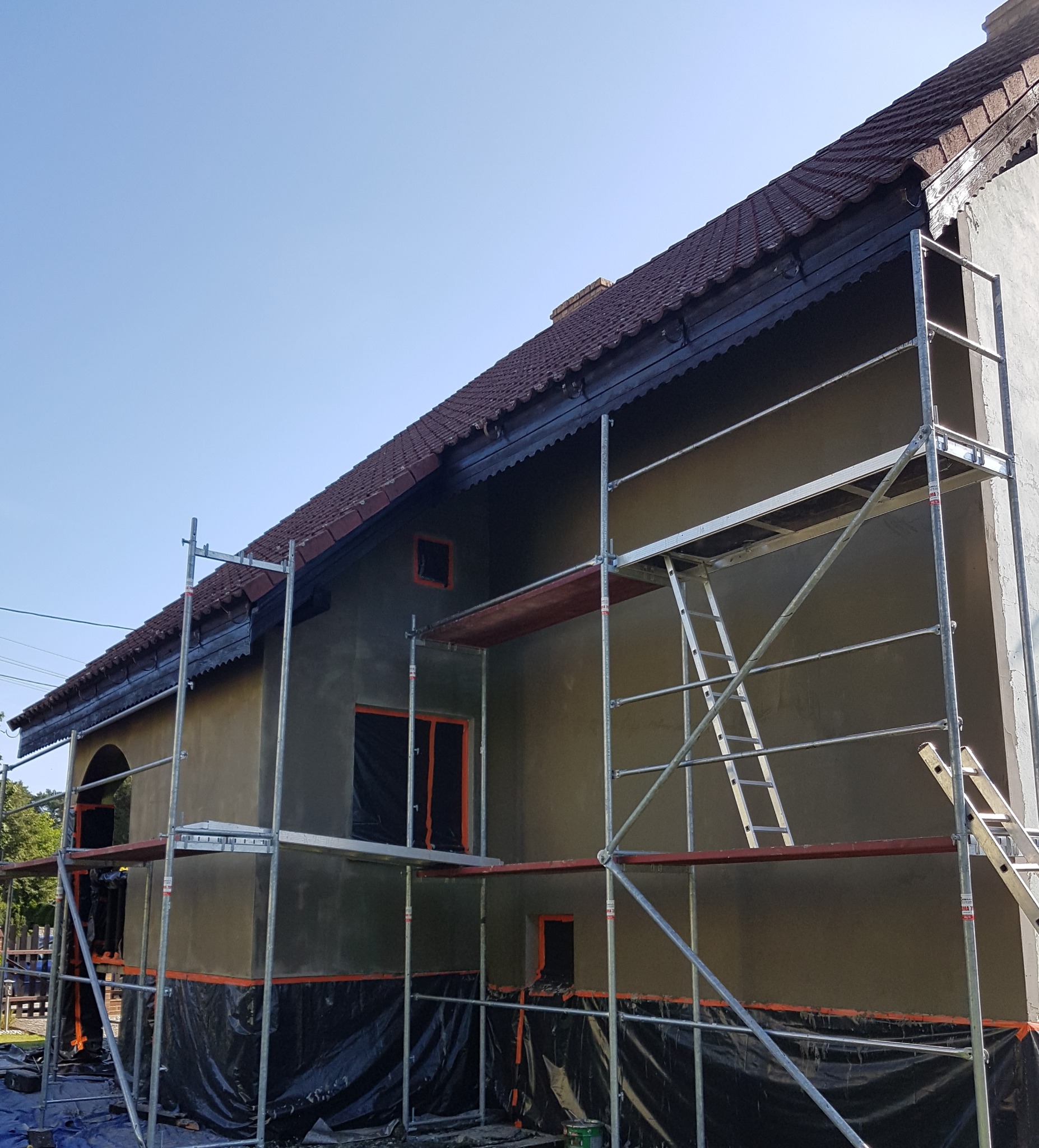 Dom jednorodzinny w trakcie prac elewacyjnych, z widocznymi rusztowaniami, zabezpieczeniem okien folią i świeżo nałożonym tynkiem.