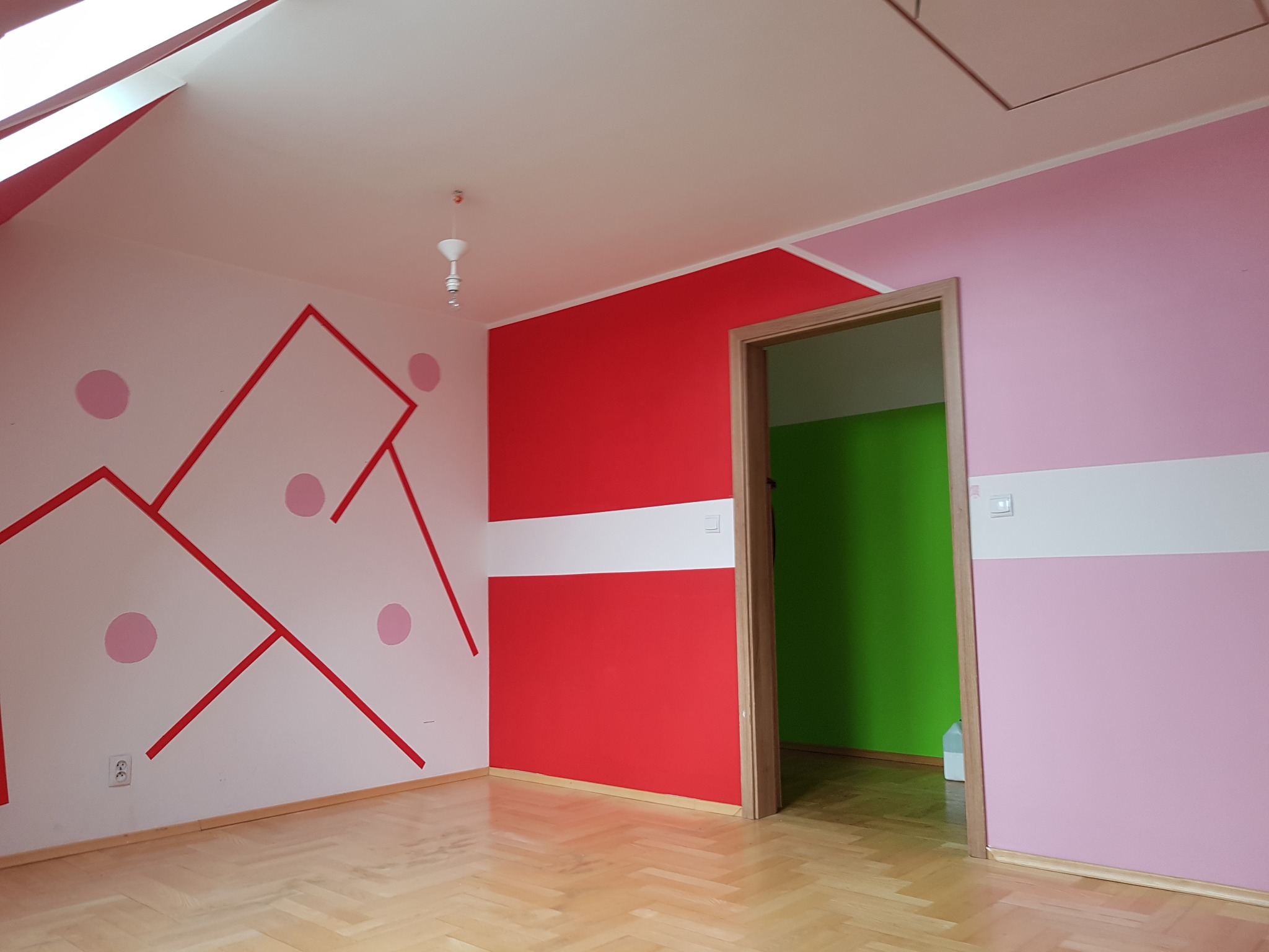 Pokój dziecięcy po malowaniu ścian w odcieniach różu, czerwieni i bieli, z geometrycznymi wzorami i okrągłymi naklejkami, parkiet na podłodze, widoczne okno dachowe.